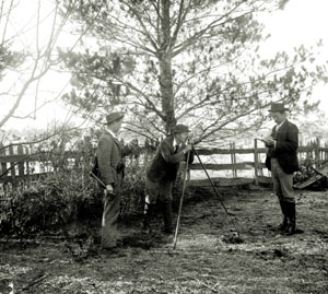 Three men standing in a field using a surveying tool on top of a tripod to examine the landscape