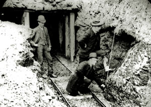 Two men inspecting the entrance to a mine with a railway track coming out of the opening