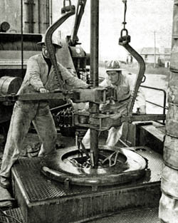 Black and white photo of two men turning a large, metal mechanical drill into the ground