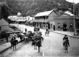 A black and white photo of seven men sitting on horses in the main street in Walhalla with loaded packhorses